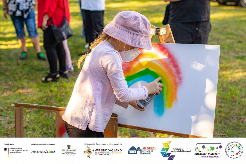 Ein Mädchen mit Maske sprüht einen Regenbogen auf eine Leinwand