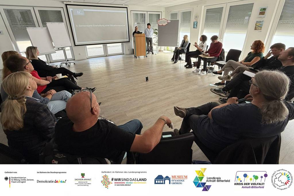 Ein Veranstaltungssaal: Etwa 15 Personen sitzen in einem Stuhlkreis und schauen nach vor. Dort stehen an einem Rednerpult zwei weitere Personen